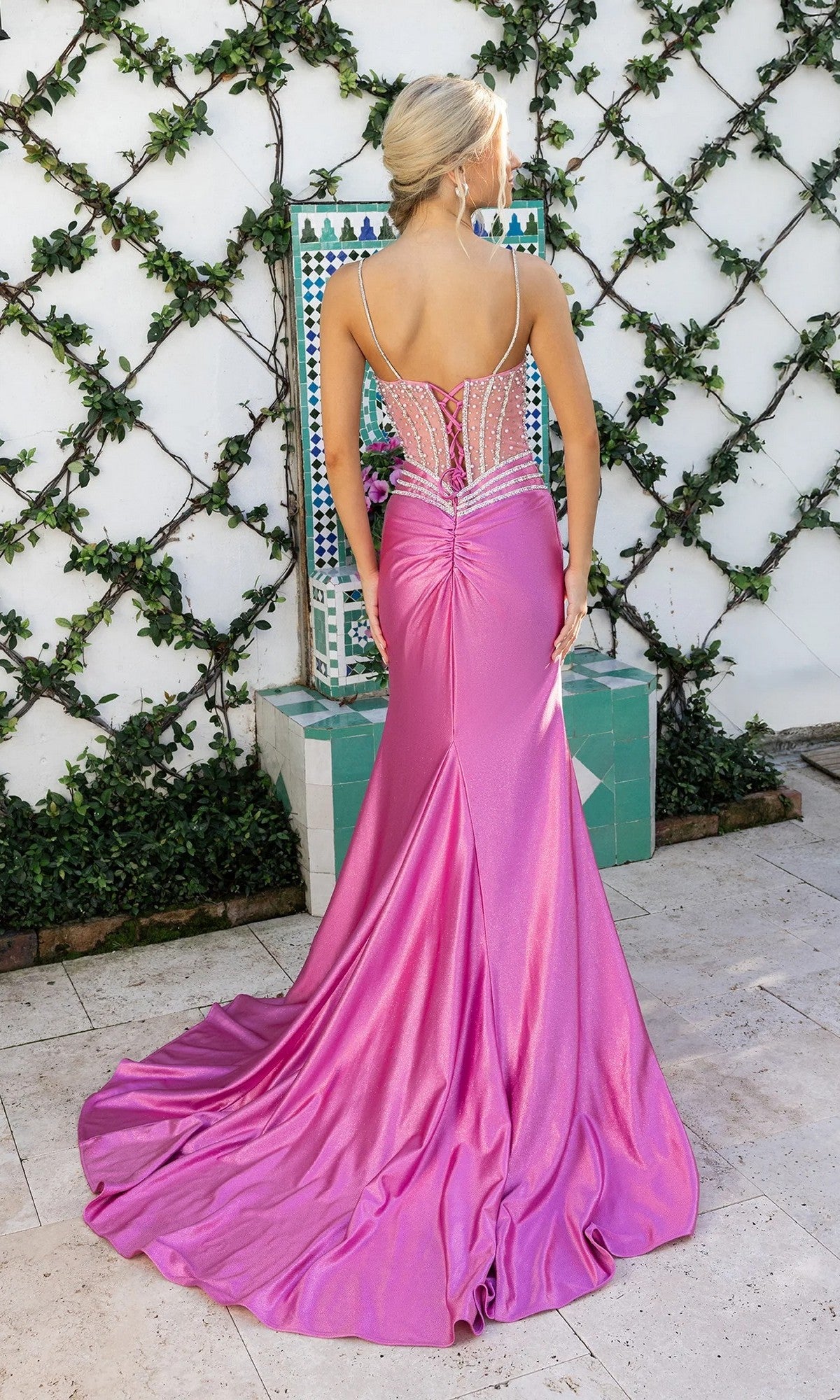 A woman in the Portia and Scarlett XO157 long formal dress, featuring a pink crystal-embellished bodice and beaded sheer back, stands outdoors by a tiled fountain and vine-covered wall.